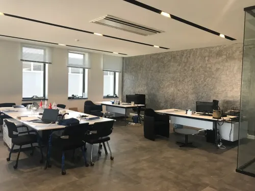 Birden fazla çalışma masasının yer aldığı ofis alanı, gri zemin ve sade mobilya diliyle kurumsal çalışma düzenini dengeli biçimde destekler.