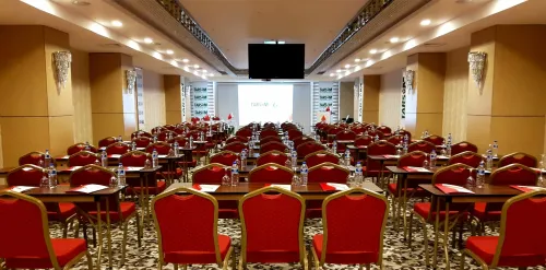 Geniş etkinlik salonu, kırmızı sandalyeler ve sıralı masa kurgusuyla otel toplantıları için temsil gücü yüksek bir kullanım alanı oluşturur.