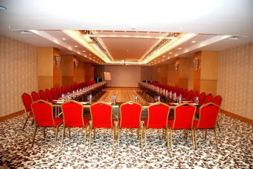 Geniş konferans salonu, kırmızı sandalye sıraları ve merkezi masa düzeniyle büyük ölçekli otel etkinlikleri için güçlü bir mekân kurgusu oluşturur.