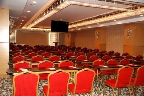 Sıralı masa ve kırmızı sandalye yerleşimiyle konferans salonu, sunum, eğitim ve otel içi kurumsal etkinlikler için düzenli bir alan sunar.