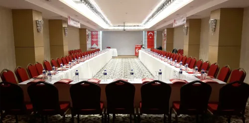 U düzeninde hazırlanan uzun masa yerleşimi, kırmızı sandalyeler ve sunum aksıyla otel toplantı salonuna kurumsal bir kullanım dili kazandırır.